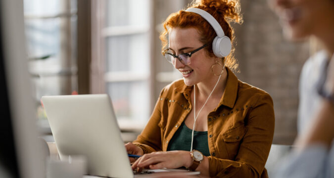 Happy red headed woman looking at her laptop and wearing headphones